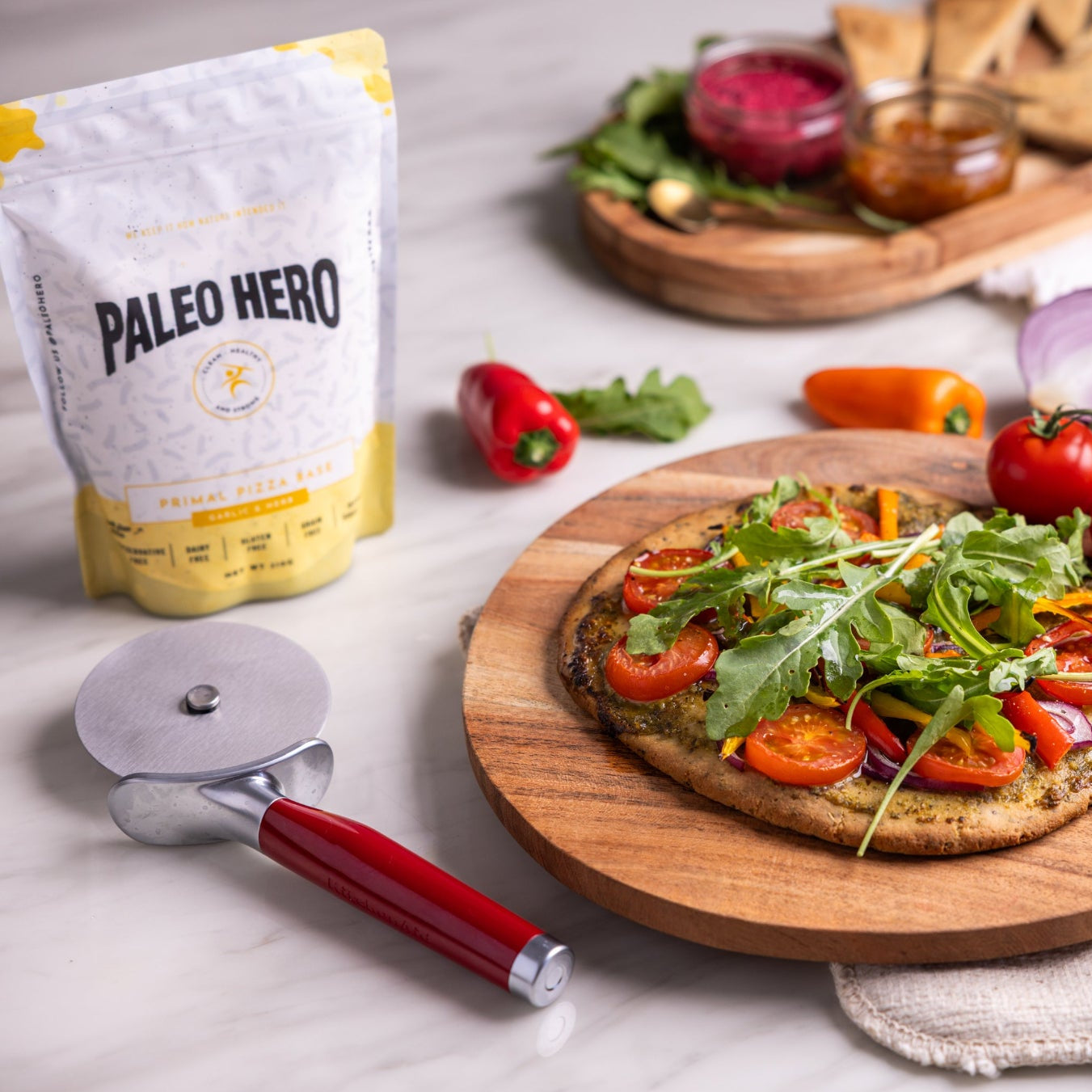 Vegetable pizza on a wooden board with Paleo Hero packaging and pizza cutter.