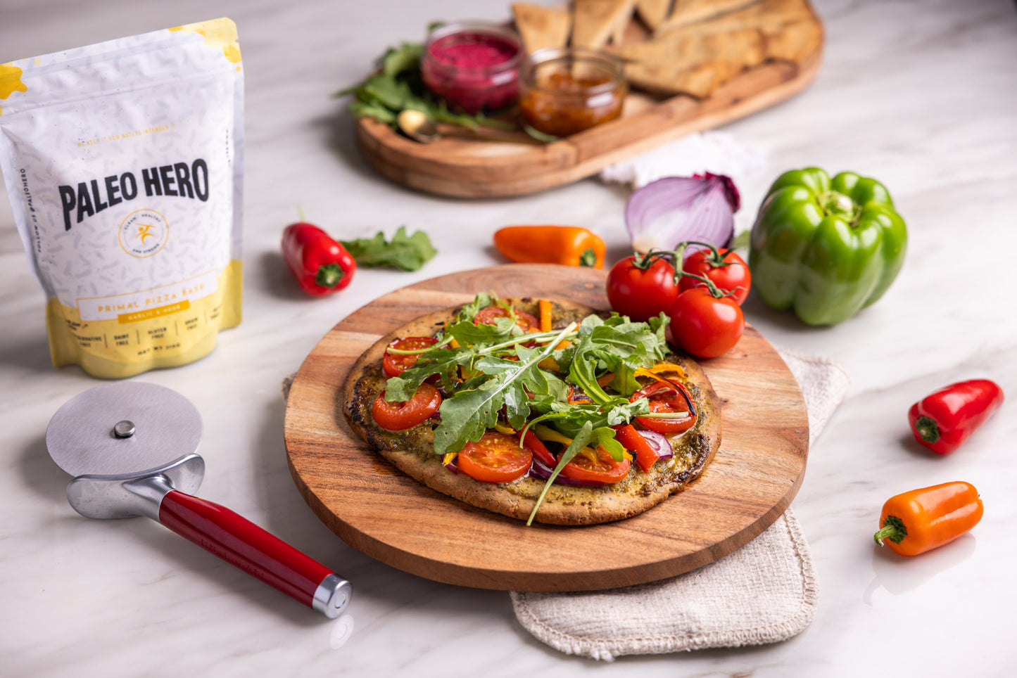 Vegetable pizza on a wooden board with Paleo Hero packaging and pizza cutter.