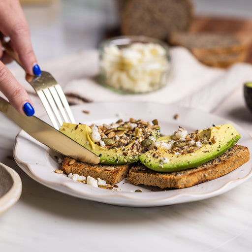 Person spreading avocado on toast with a knife, surrounded by ingredients on a table.