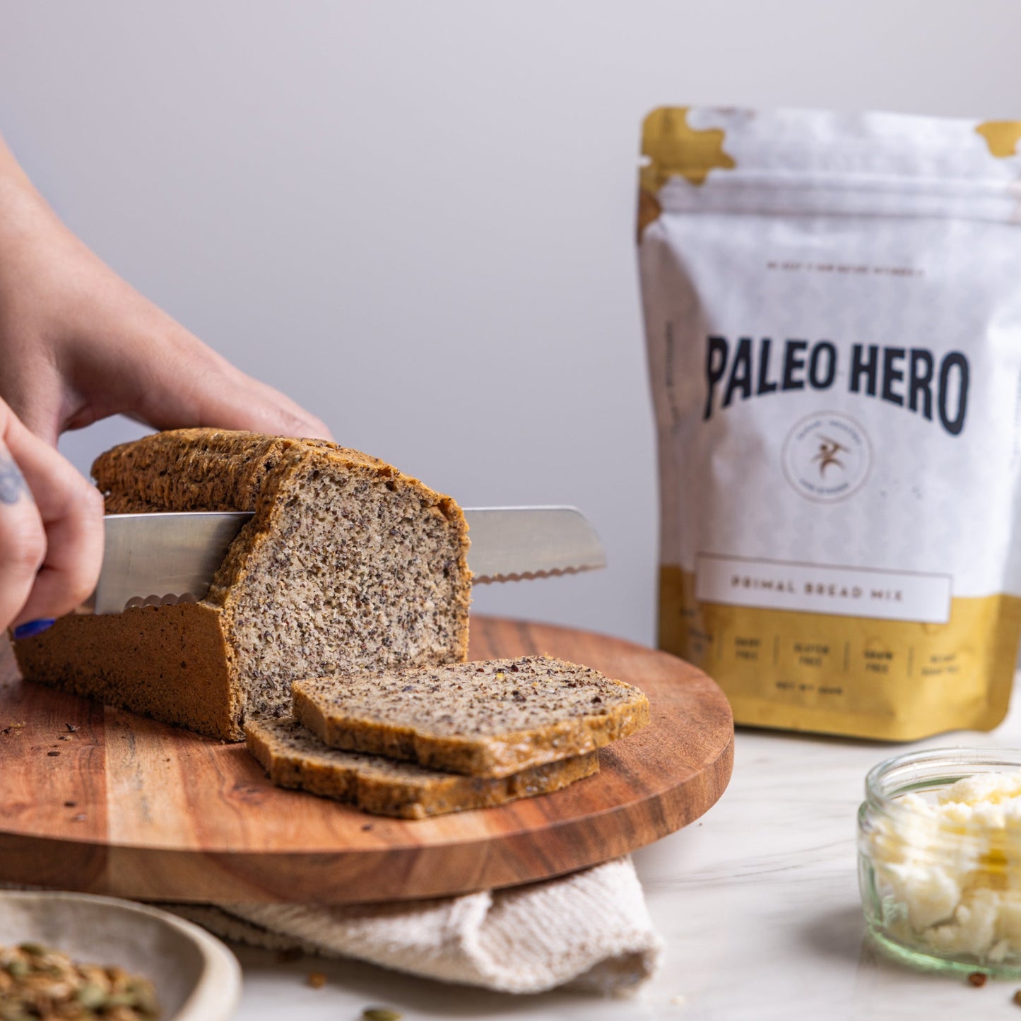 Person slicing bread on a wooden board with a 'Paleo Hero' package in the background.