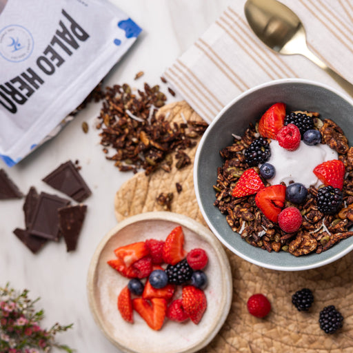 Bowls of granola with fruits and a package labeled 'Paleo Hero' on a marble surface.