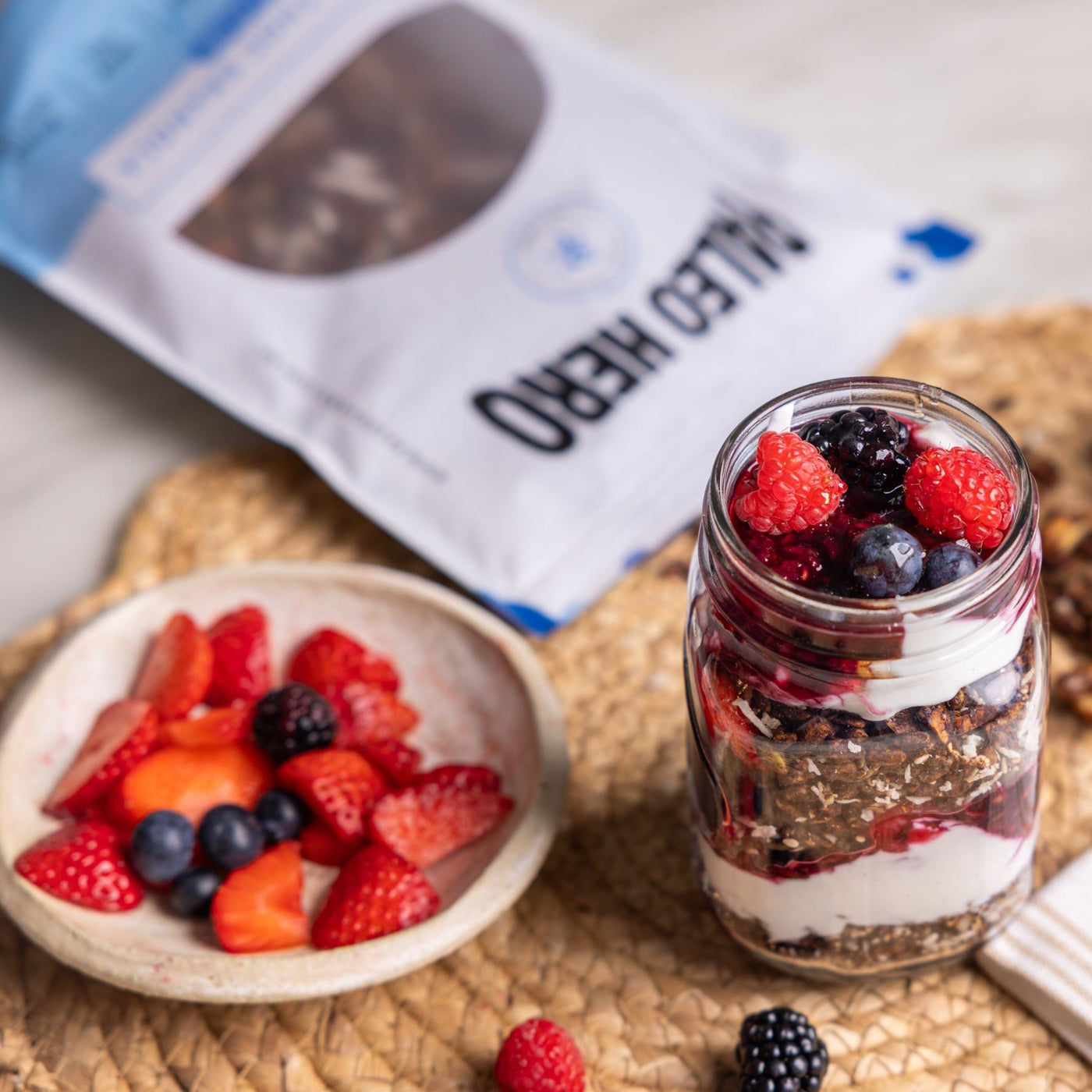 Mason jar with layered dessert, berries, and honey on a woven mat with bag of Paleo Hero Chocolate Muesli on its side