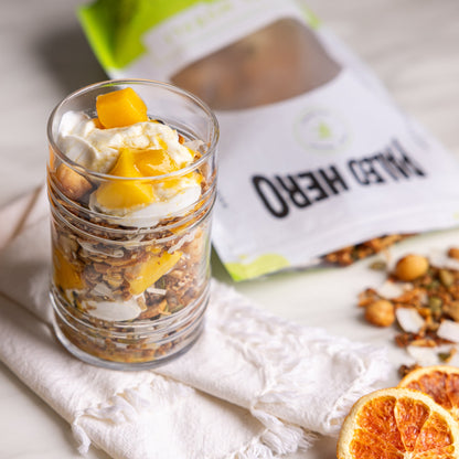 Glass of yogurt with granola and fruit on a white cloth with a spoon and 'Whey Hero' package in the background.