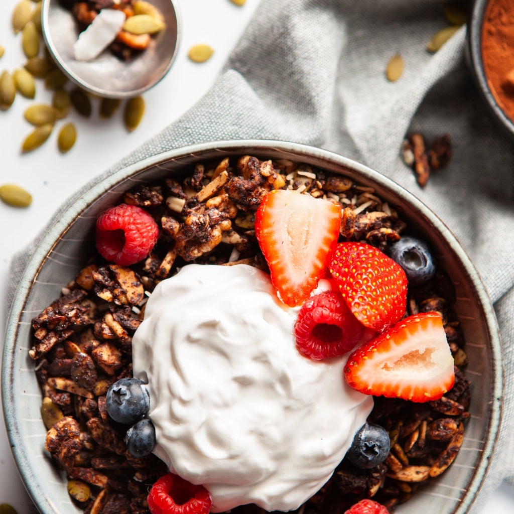 a bowl of chocolate granola with strawberries, blueberries, a dollop of cream, on a light background